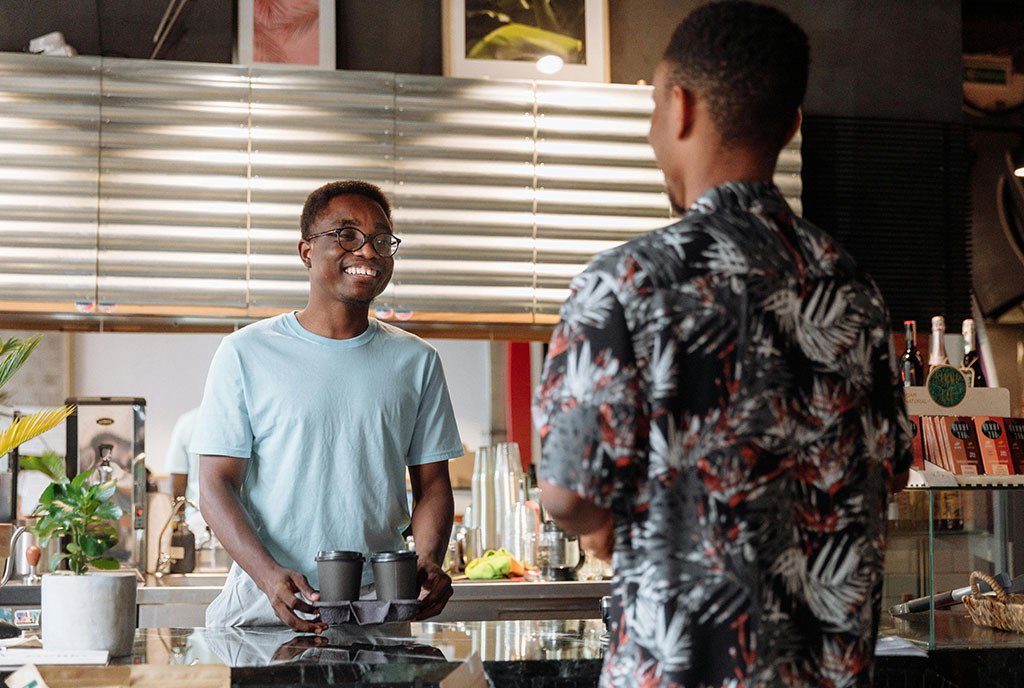 A Black man shopping at a fellow Black man’s coffee shop, as the owner smiles and hands him an order of two coffees