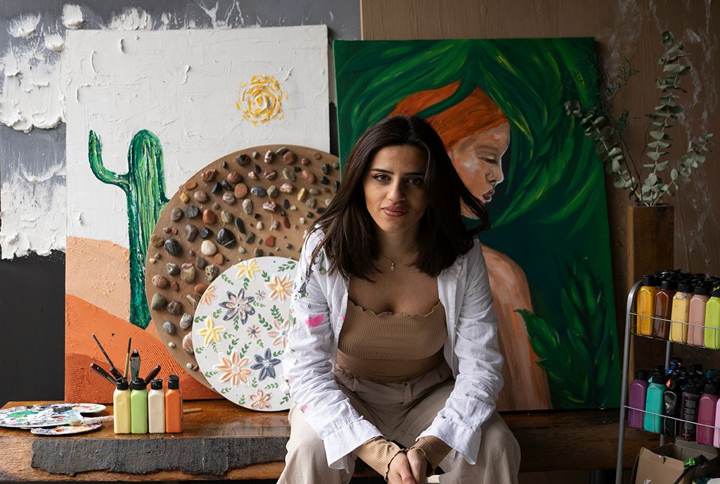 A brown woman leans forward with a content smile, while sitting in front of her painting.