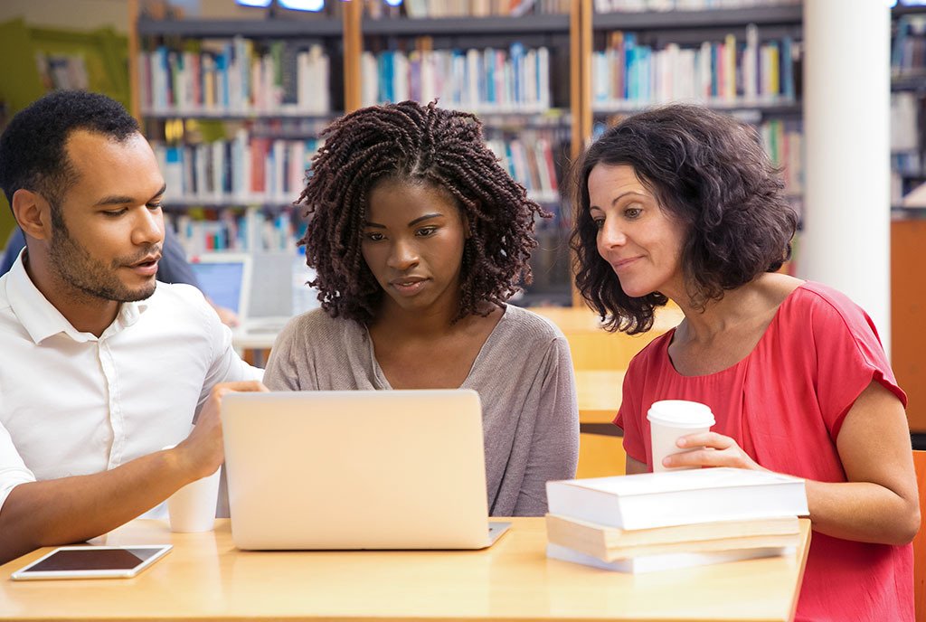 A Black woman with short twists looking at a computer screen in an adult learning class hosted in a library. She is sandwiched by a man and woman who are assisting her.