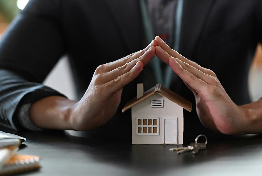 An insurance agent with their fingers pressed together over the roof of a small model home, as if protecting it.