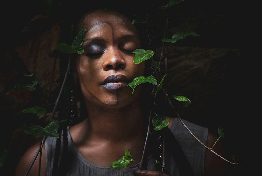 A woman with her eyes closed, nestled beneath a vine of leaves. Half of her face is made up with purple lipstick and eye shadow, the other side is natural.