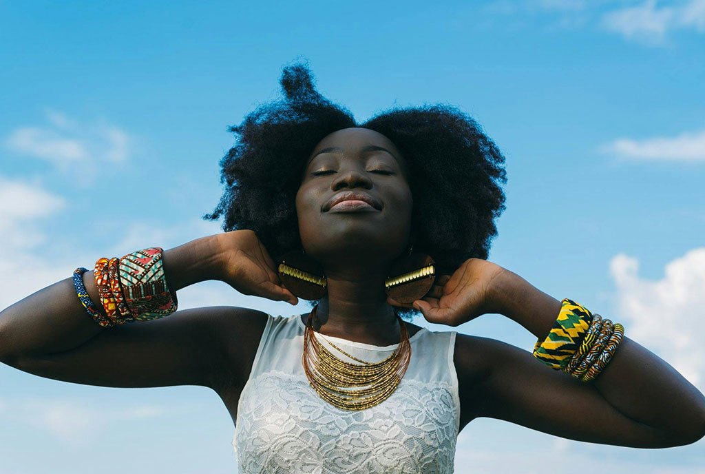 A Black woman with 4c hair smiling to herself as she closes her eyes and cradles the back of her neck.