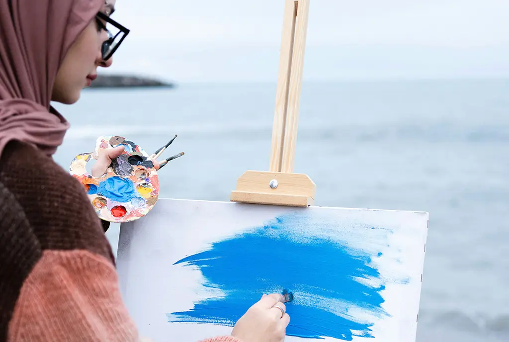 A woman painting on a canvas in front of a natural body of water, using art as a refuge and connection to the environment.