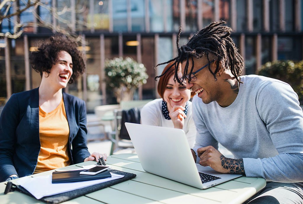 A group of diverse co-workers laughing and talking together as they gather around a computer screen.