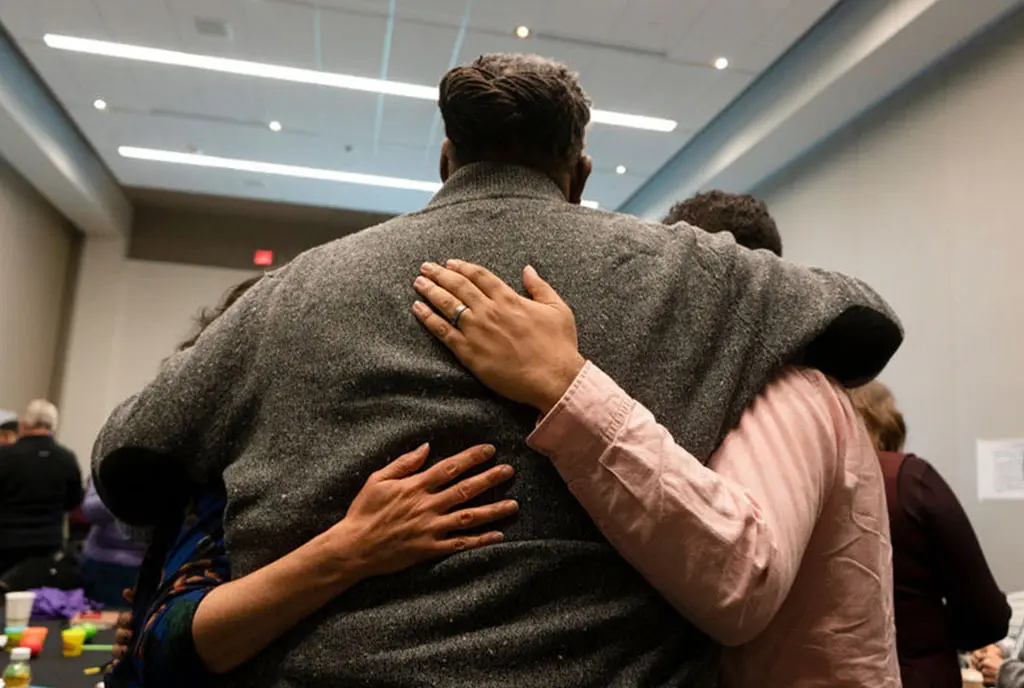 Facing Race participants embrace each other in a conference room, with the arms of two participants around the back of a Black man with locs.