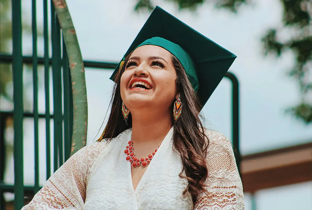 A Native American college graduate wearing a green graduation cap, smiling and looking up hopefully.