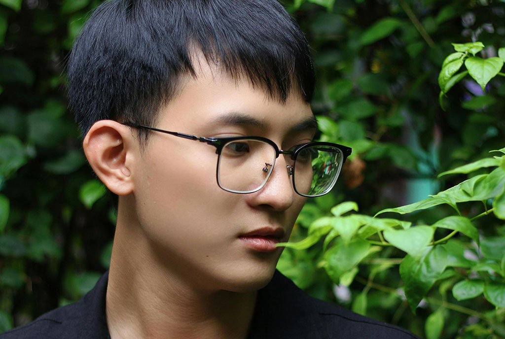 A young Vietnamese man in a Black shirt and eyeglasses standing near a plant, and looking down with a concerned look