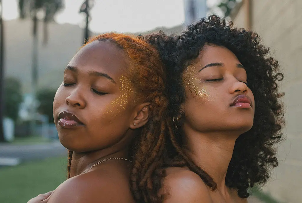 Black women who are leaning against each other with their eyes closed. They have relaxed, but tired expressions on their faces.