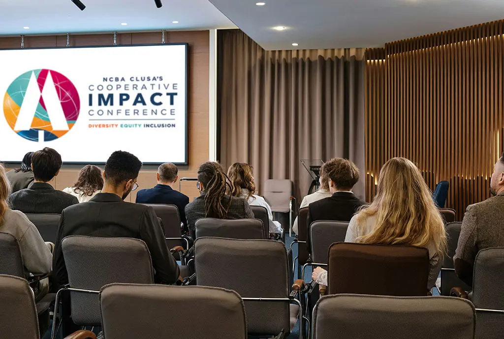 A group of diverse people sitting in a break-out room during a conference. The NCBA CLUSA Cooperative Impact Conference logo is on a projector screen at the front of the room.