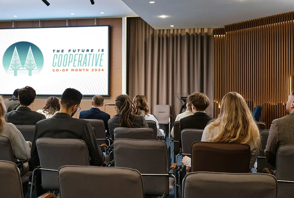 A group of diverse people sitting in a break-out room during a conference. “The Future is Cooperative” logo is on a projector screen at the front of the room.