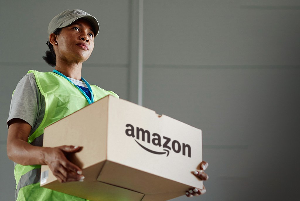 A Black woman holds a cardboard Amazon box in her hands, as she looks off into the distance with a serious look on her face.