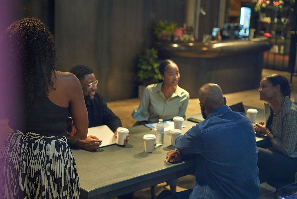 A team of Black tech workers collaborating around a table. Their computers and devices are visible as they speak with each other