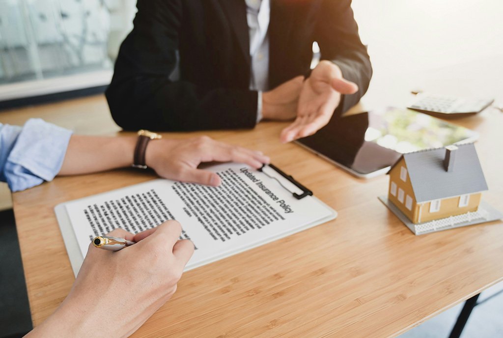 A person signing an updated insurance policy, as an insurance agent gestures. There is a small model house and calculator in the background.