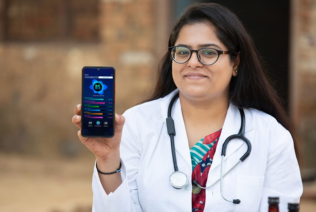 Portrait of a young smiling Indian female doctor, standing in front of a rural clinic and wearing a stethoscope while holding up a phone with a health app on the screen.