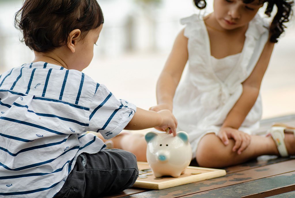 Two small children placing coins into the back of a piggy bank, symbolizing gradual wealth-building over time.