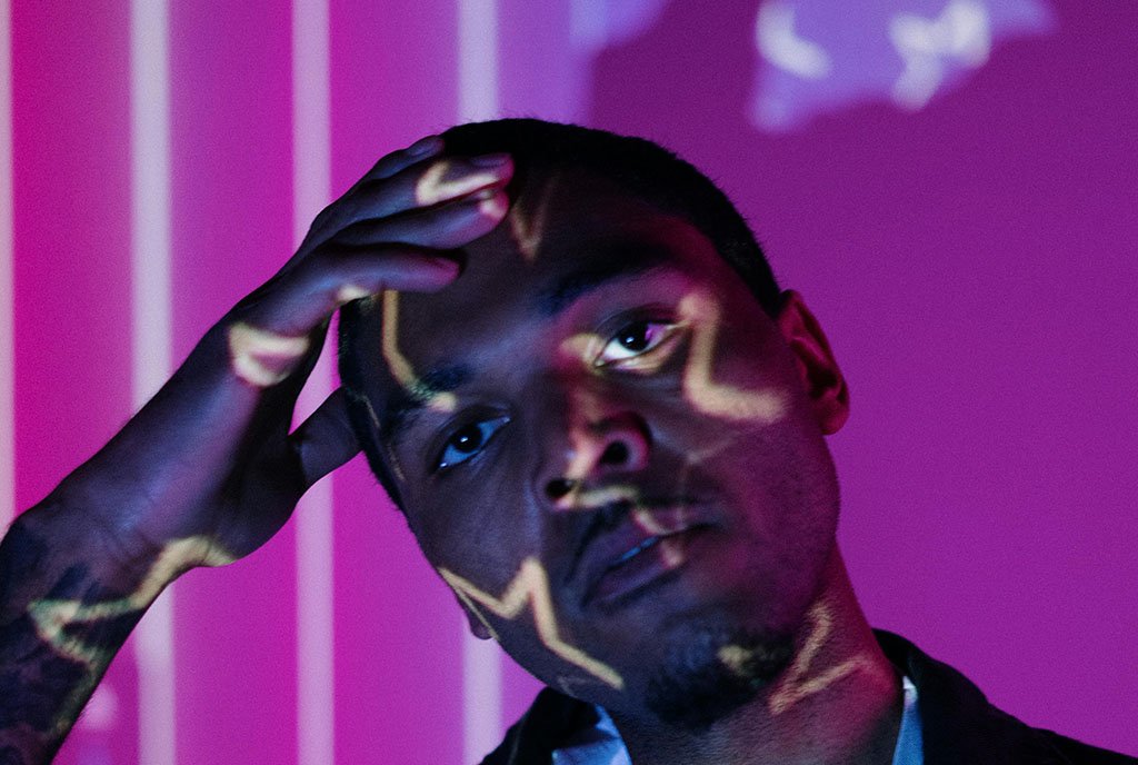 A powerful portrait of a Black man standing in front of an abstract projection of the American flag, symbolizing systemic critique.