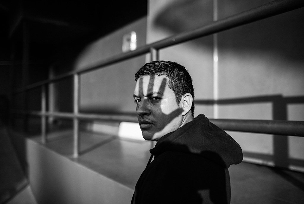 A Black and White photograph of Indigenous man looking over his shoulder into the camera with his face covered by the shadow of a hand, symbolizing silencing and exclusion of Native voices in climate spaces.