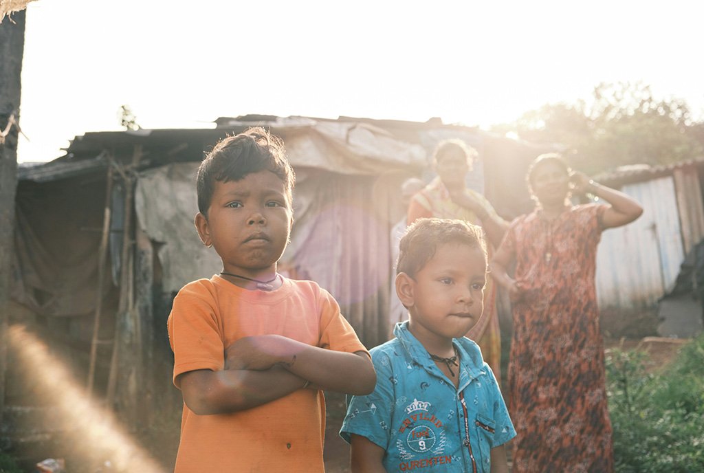 Two young boys stand outside of their home in the slums, with two women watching from behind. The sun’s rays glitter behind and around them.