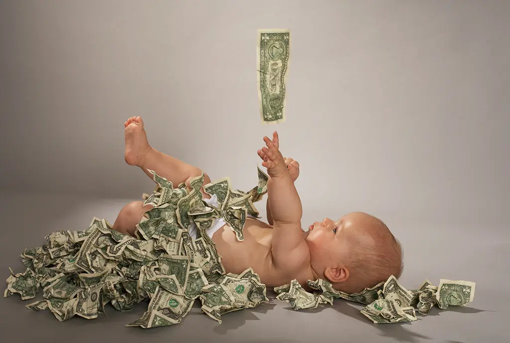 A newborn baby cradled in a bed of dollars bills, as he reaches up for one dangling above his head, representing financial growth and stability from baby bonds.