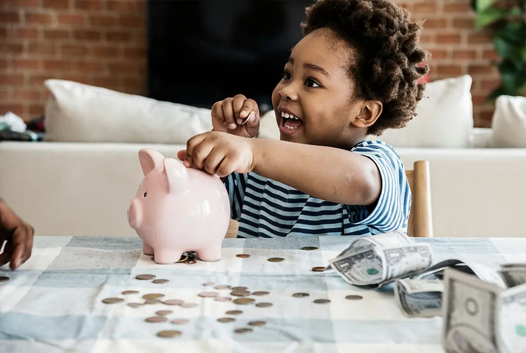 A young Black toddler boy, with a textured afro smiling as he puts coins into a pink piggy bank, symbolizing the potential wealth growth from a young age.