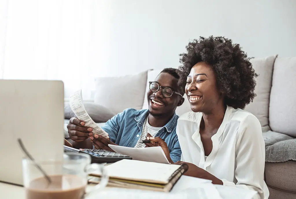 A young Black couple sitting in front of their computer and calculator reviewing receipts and paying bills, suggesting financial stability and hope.