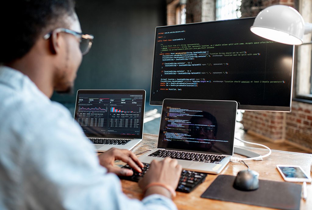 A Black man working at a desk with multiple computer screens showing complex coding