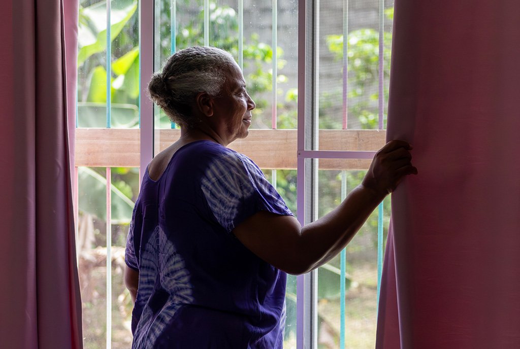 An elderly Black woman looks out of her window as she adjusts the curtain with a hopeful smile on her face.