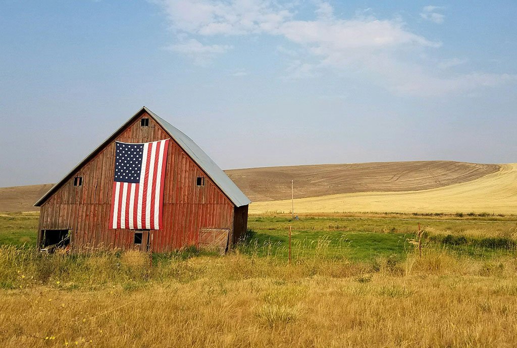 A rural barn in a Washington state grassland, with an American flag strung across the top. Washington, USA. 2024.