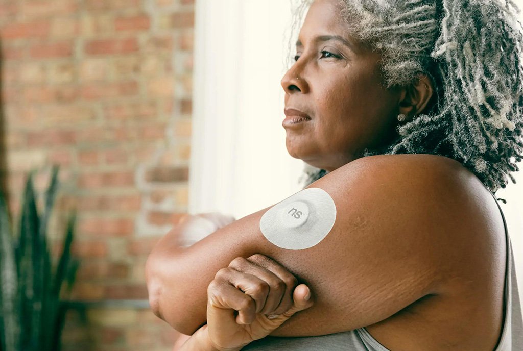 An elderly Black woman with gray locs stretching and revealing a continuous glucose monitor (CGM) on her arm).