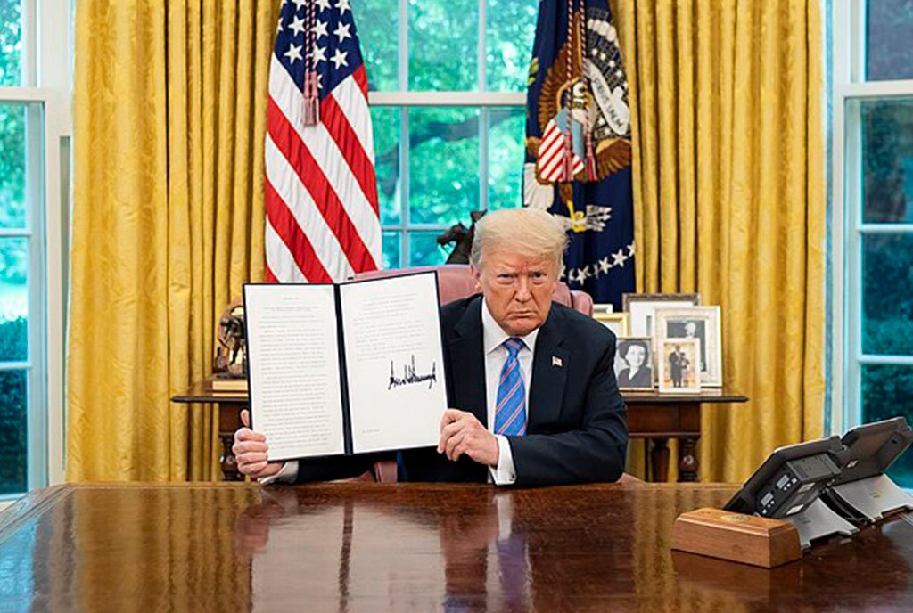 President Trump sitting in the oval office and holding up an executive order with his signature on it. He has a determined and stoic look on his face.