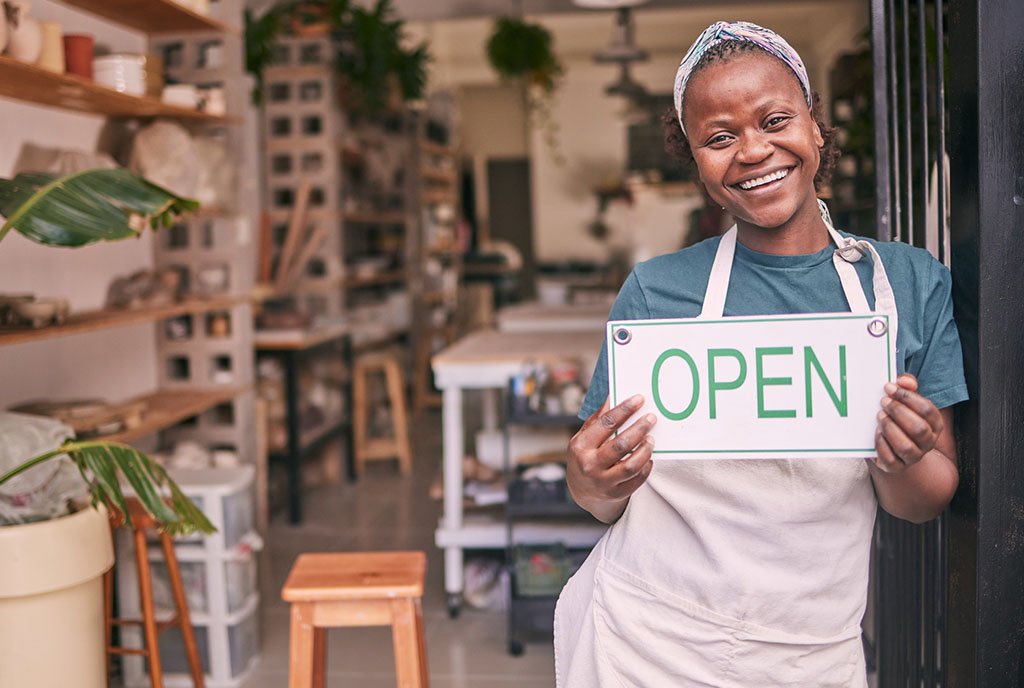 A Black woman business owner wearing an apron in her store front holding up an “Open” sign, symbolizing the potential impact of adequate start-up capital for Black business owners.