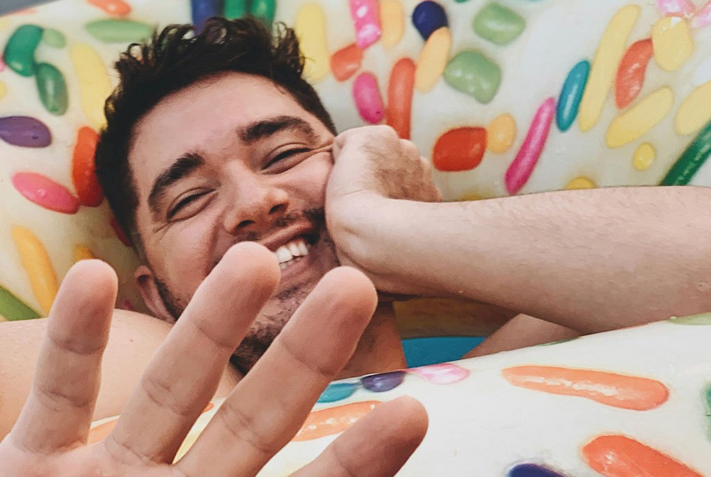 A grinning Latino man settled in a donut-shaped pool toy, looking at the camera and blocking the shot playfully with his hand, symbolizing the hopeful outlook of community-driven mental health solutions.