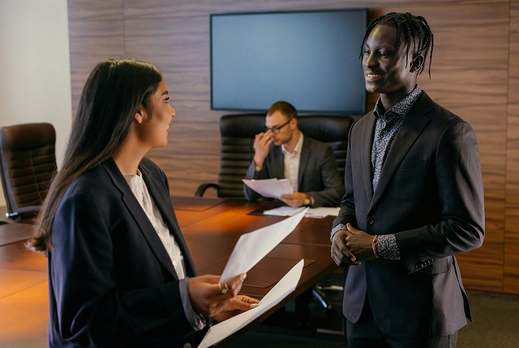 A diverse group–a Black man, a Brown woman, and a White man–happily conversing in a board room, symbolizing a liberatory board that prioritizes Loving Accountability and Abundant Resourcing.