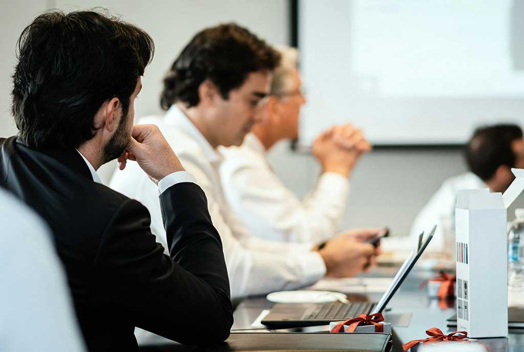 A boardroom scene showing a group of middle-aged white men sitting on one side of a conference table in suit jackets and button downs, a relic of the consolidation of wealth and influence that boards offer to elites.