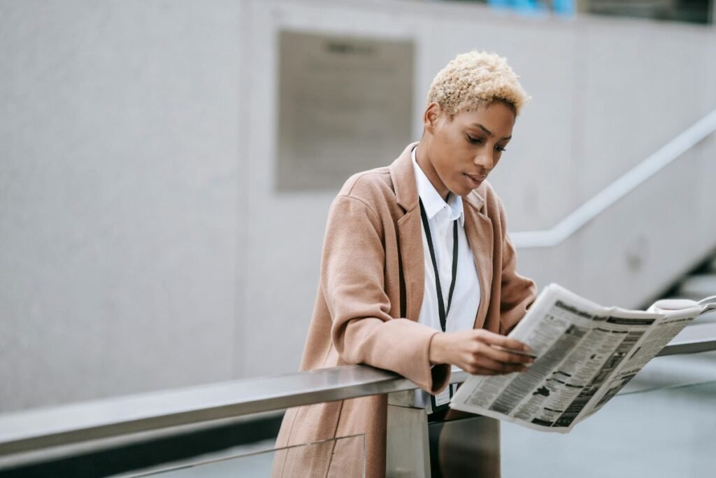 A woman with light brown skin and blond natural curly hair leans forward against a railing reading a newspaper.