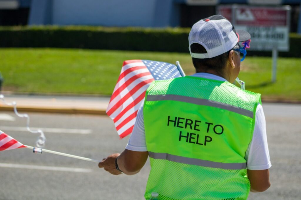 A person in a baseball cap holds two American flags and wears a bright green vest reading “Here to Help” on the back.
