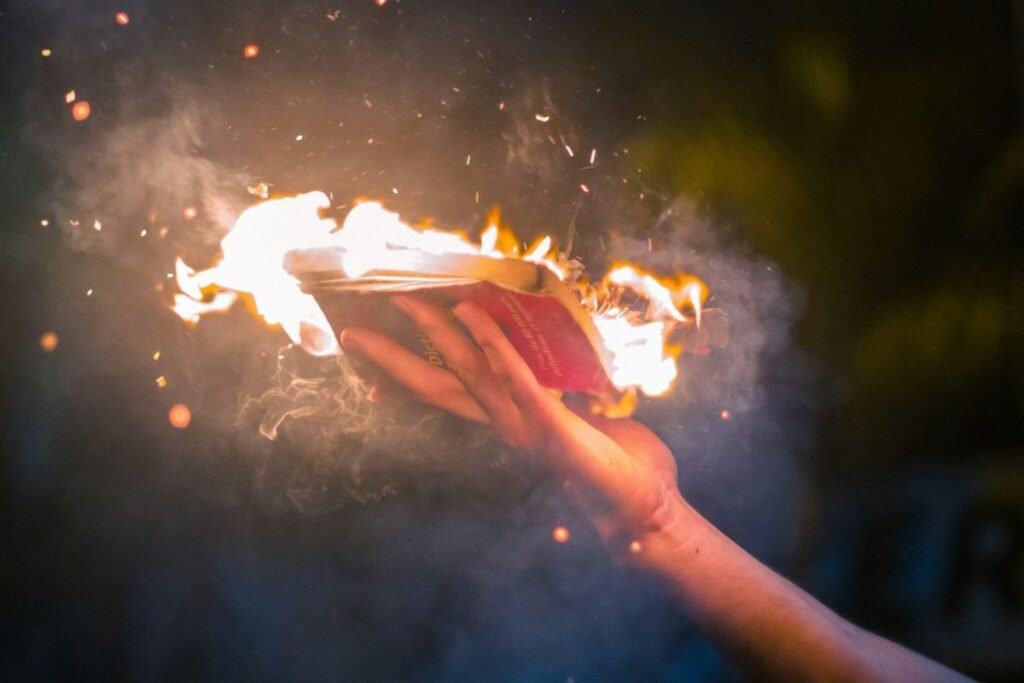 An outstretched hand holds an open paperback book with a black and red cover. Flames and sparks come from the pages of the book and smoke fills the night sky around it.