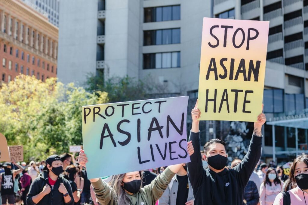 Two people wearing black face masks in a crowd of protestors hold up signs on rainbow-colored cardboard. One sign reads: Protect Asian Lives. The other: Stop Asian Hate.