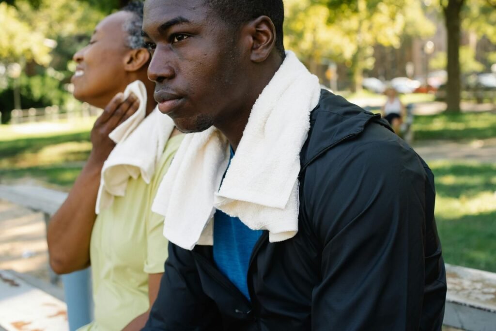 A close-up of a man with dark brown skin sitting outside in a park with a white towel around his neck. An older woman with brown skin sits next to him, wiping her neck with her towel in the heat.