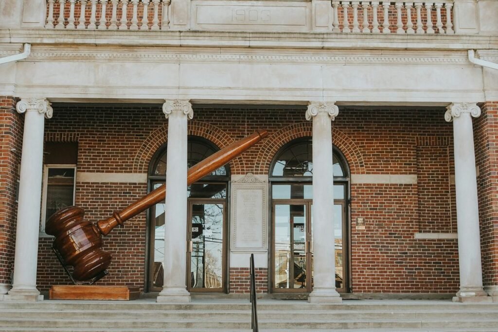 A statue of a giant wooden gavel on the porch of a brick courthouse with large front steps and white stone pillars.
