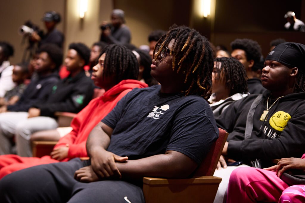 A group of young Black men sit attentively in an auditorium listen attentively during a forum.