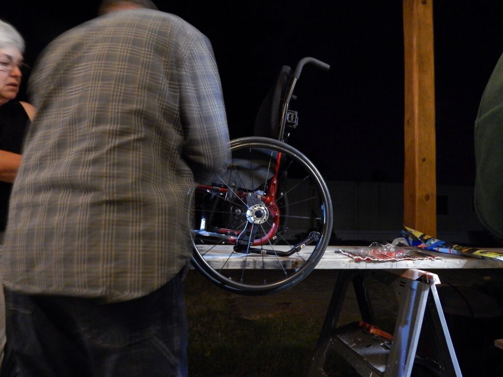 Two people repair a wheelchair on a workbench at night, with tools scattered and focused lighting on the wheel.