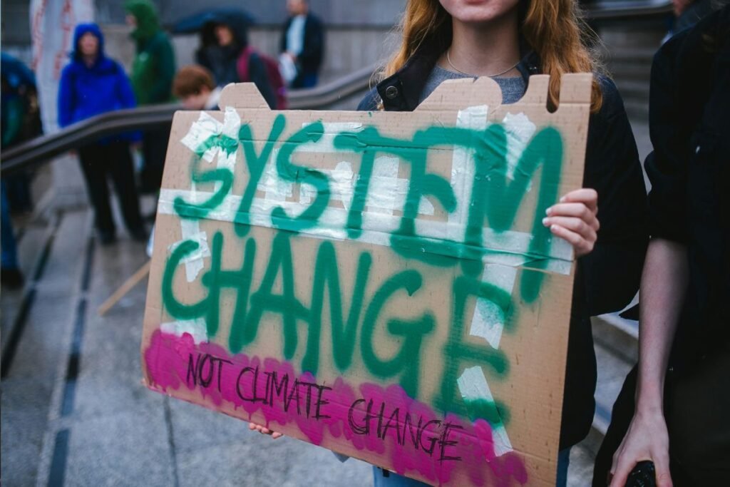 A young person with red hair holds up a cardboard sign that reads System Change, Not Climate Change at a protest.