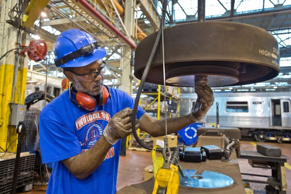 A maintenance worker wearing a union t-shirt inspects subway car parts at Coney Island Yard, an industrial facility.