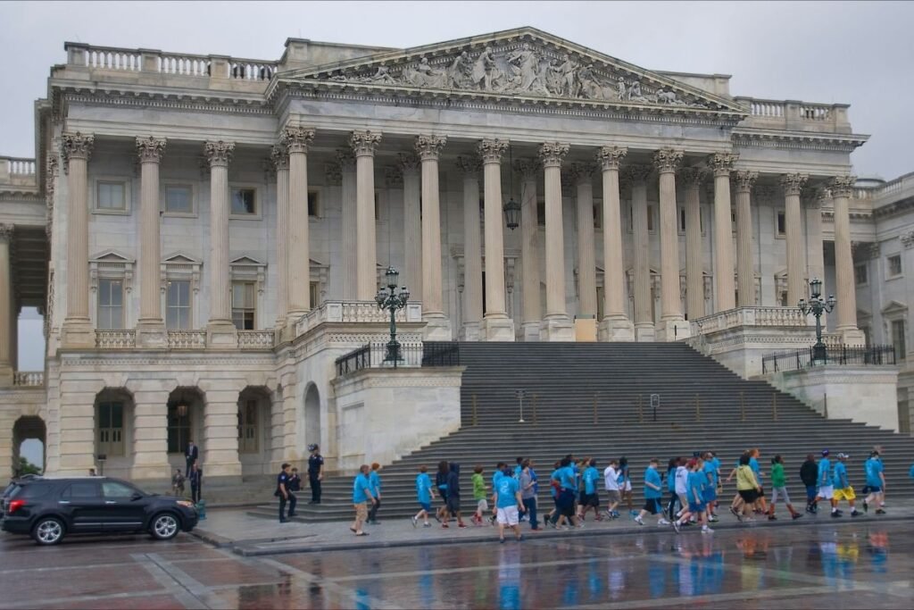 A group of about two dozen students, many wearing blue shirts, walk in the rain in front of the US House of Representatives.