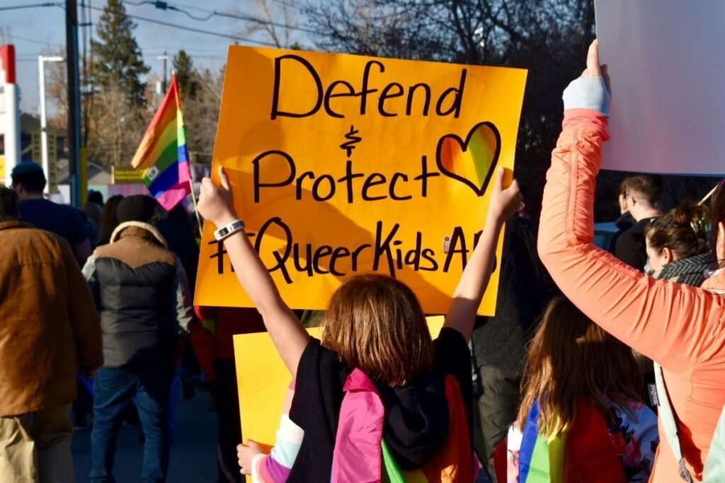 A child holds up a sign at a Pride gathering that reads Defend + Protect Queer Kids.