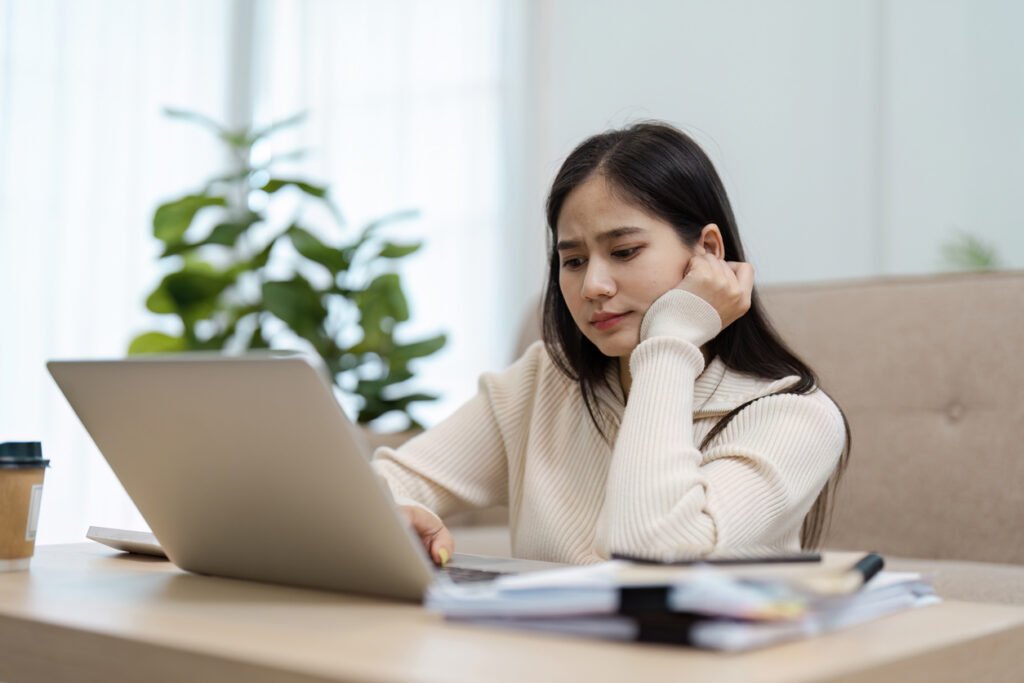 A thoughtful young Asian woman sits at her table, appearing concerned as she navigates challenges on her laptop at home.