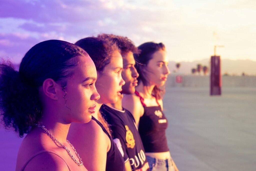 Four young people stand in a parking lot, looking at the sunset. There are mountains and palm trees in the distance.