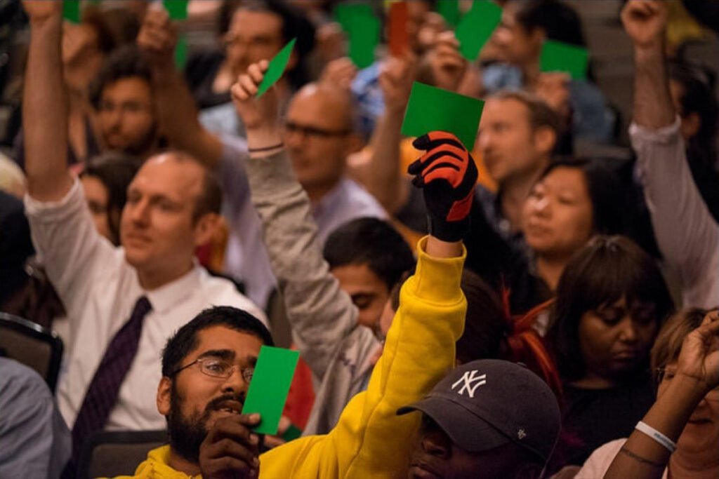 A dozen or so people who are part of a larger crowd lift up green cards to vote in favor of a proposal at a public meeting.