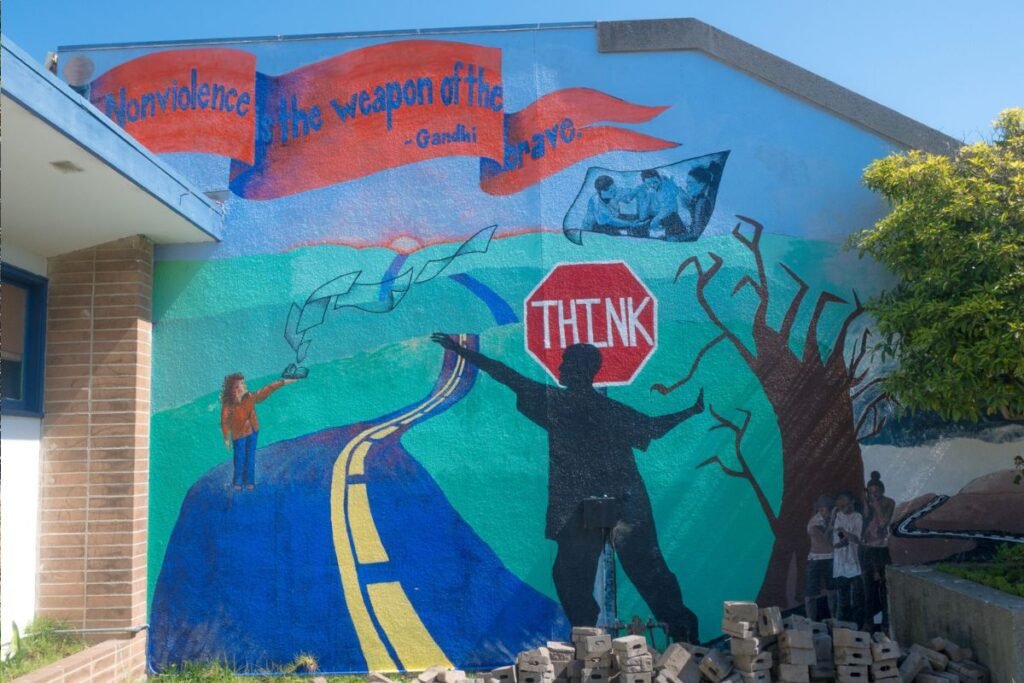 A mural on war and peace: A sign says “think” at start of blue road. A woman flies a kite. A Ghandi saying is on a banner.